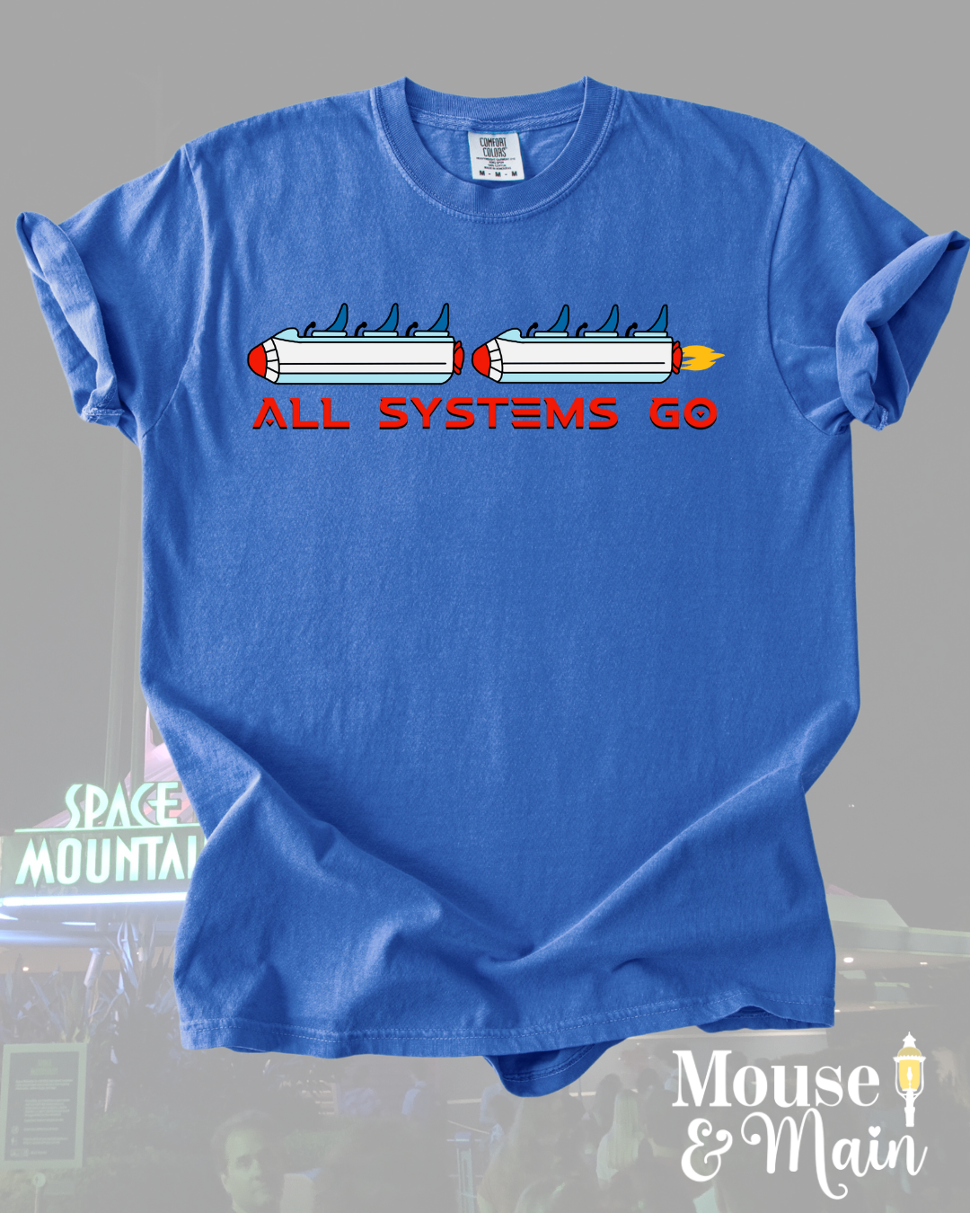 Tomorrowland-inspired graphic tee with rocket ride design and bold space-age font, displayed flat on a gray background with subtle park imagery.